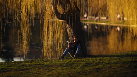 Junger-Mann-Liest-Ein-Buch-Und-Sitzt-In-Der-Abenddämmerung-Unter-Einem-Goldenen-Baum-An-Einem-öffentlichen-See-Im-Stadtpark