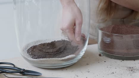A-young-female-botanist-creates-a-tiny-live-forest-ecosystem-in-a-huge-glass-jar---putting-the-decorating-sand---a-tight-close-up