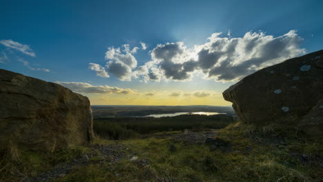 Time-lapse-of-rural-farming-landscape-with-lake,-forest-and-hills-during-a-cloudy-sunset-viewed-between-large-rocks-from-above-Lough-Meelagh-in-county-Roscommon-in-Ireland