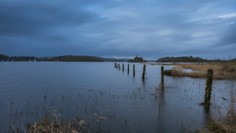 Timelapse-of-local-water-sport-yachting-boats-with-castle-ruin-in-distance-and-old-fence-pillars-in-foreground-at-Lough-Key-in-county-Roscommon-in-Ireland-on-a-cloudy-day