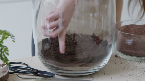 A-young-female-botanist-creates-a-tiny-live-forest-ecosystem-in-a-huge-glass-jar---finishing-the-soil-layer---a-tight-close-up