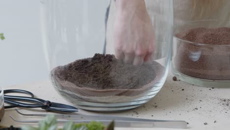A-young-female-botanist-creates-a-tiny-live-forest-ecosystem-in-a-huge-glass-jar---leveling-the-main-soil-layer---a-tight-close-up