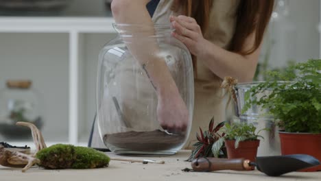 A-young-female-florist-prepares-the-first-layer-of-soil-in-a-glass-jar-for-creating-a-tiny-live-forest-ecosystem---close-up