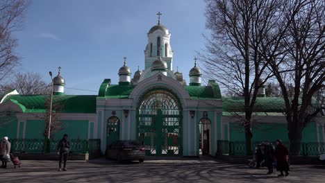 People-walk-past-a-Christian-Orthodox-church-with-a-green-gate,-roof-and-walls-and-golden-domes