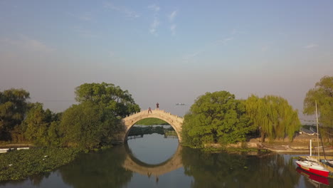 Stone-arch-bridge-forms-circle-reflected-in-weed-choked-lake,-aerial