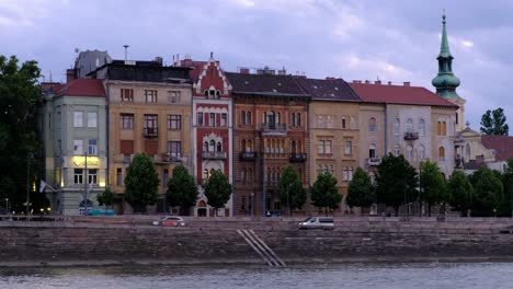 Danube-river-cruise,-church-spire-and-building-facades-during-golden-hour,-beautiful-cotton-candy-clouds,-house-lights,-cars-driving-on-road,-coast-line,-river-side,-embankment,-tourists-sitting