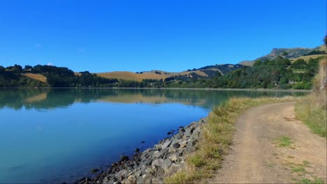 Walking-on-coastal-track-in-harbor-with-nice-reflection-of-low-hills-on-calm-water---Governors-Bay-Track,-Banks-Peninsula