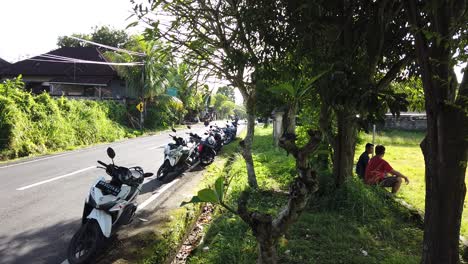 Zwei-Freunde-Unterhalten-Sich-An-Einem-Sonnigen-Tag-In-Gianyar-Ubud-Neben-Einer-Balinesischen-Dorfstraße-Mit-Motorrollern,-Die-In-Der-Nähe-Grüner-Felder-Fahren