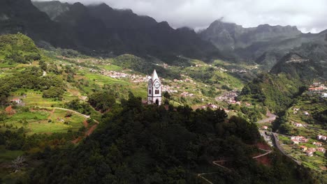 Aerial-reveal-of-old-chapel-on-top-of-green-hill-with-mountain-background-landscape,-Sao-Vicente,-Madeira