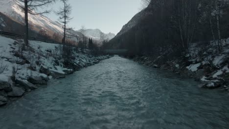 Dolly-over-cold-Swiss-flowing-river-water-with-snow-on-river-banks-in-Switzerland-Alps-with-epic-mountain-panorama