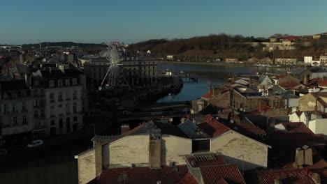 Drone-flying-over-buildings-rooftops-with-ferris-wheel-along-river-shore,-Bayonne-in-France