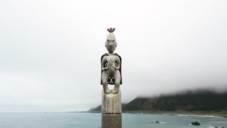 Aerial-pan-shot-of-a-traditional-Māori-statue-at-the-top-of-a-totem-pole-in-front-of-large-bay-in-New-Zealand-on-a-cloudy-day