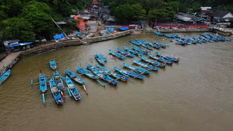 Orbit-drone-shot-of-many-fishermen-boats-anchored-on-the-harbour-with-market-buildings-and-fish-auctions---Baron-Beach,-Yogyakarta,-Indonesia