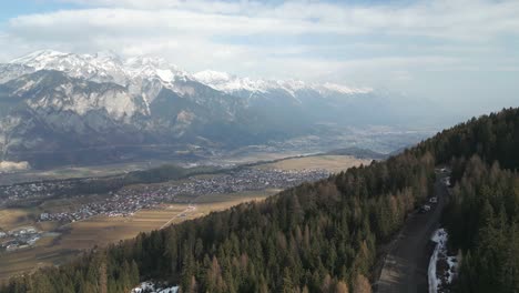 Aerial-drone-flying-over-forests-with-a-road-with-a-car-driving-and-snow-covered-alpine-mountain-range-and-a-city-in-the-distance-on-a-warm-and-beautiful-sunny-blue-sky-winter-day