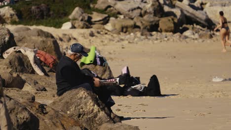 Ein-älterer-Mann-Sitzt-Auf-Einem-Felsen-An-Einem-Sonnigen-Strand-Und-Liest-Eine-Zeitung