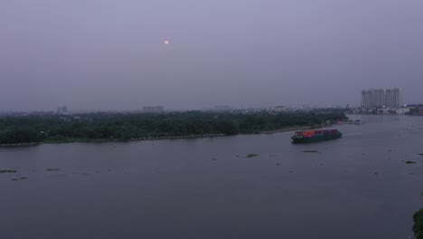 Container-boat-carrying-cargo-on-Saigon-River,-Vietnam-at-sunset-with-view-of-water-and-full-moon