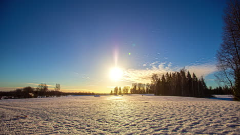 Eine-Wunderschöne-Aufgehende-Sonne,-Die-Durch-Einen-Klaren-Blauen-Himmel-Mit-Kleinen-Weißen-Wolken-über-Einem-Schneebedeckten-Feld-Mit-Einem-Wald-Im-Hintergrund-Vorbeigleitet