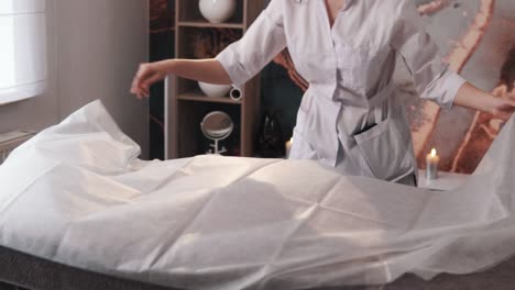 female-massage-therapist-in-a-white-robe-covers-the-massage-table-with-a-disposable-sheet