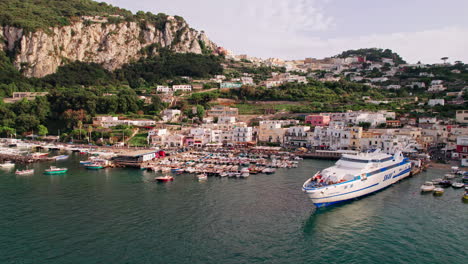 Una-Amplia-Toma-De-Drones-Que-Rodea-Un-Gran-Ferry-En-Marina-Grande-En-La-Isla-De-Capri-En-Campania,-Italia,-Con-Barcos-Y-Turistas-En-El-Puerto-Y-Un-Exuberante-Telón-De-Fondo-De-La-Isla,-Acantilados-Rocosos-Y-Edificios-De-Color-Pastel