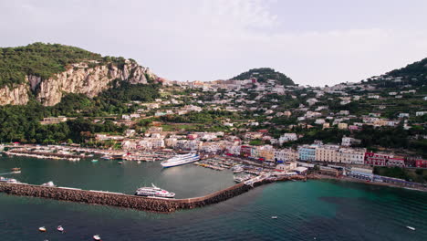 An-extreme-wide-drone-shot-approaching-Marina-Grande-on-the-island-of-Capri-in-Campania,-Italy,-showing-boats-and-an-SNAV-hydrofoil-ferry-in-the-port-as-tourists-walk-by-colorful-pastel-buildings