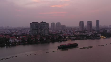 Container-boat-carrying-cargo-and-big-panning-shot-on-Saigon-River,-Vietnam-at-sunset-with-view-of-water-and-city-skyline