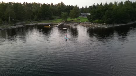 Aerial-of-man-paddle-boarding-toward-summer-lakehouse,-Hällingsjö,-Sweden