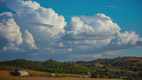 Blue-cloud-with-big-white-clouds-moving-in-a-timelapse,-over-a-meadow-with-trees-and-hills