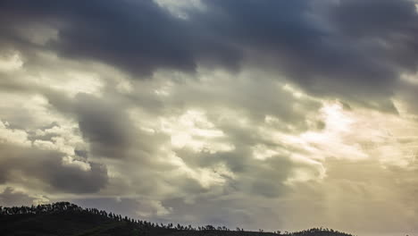 Sky-with-white-clouds-advancing-in-the-sky-and-the-rays-of-the-sun-passing-through-them-over-a-hill-with-trees