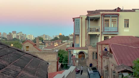 Moon-rise-over-Tbilisi-Georgia-in-the-high-mountain-of-Caucasus-in-Europe-city-center-café-historical-building-old-house-with-red-tile-roof-pink-blue-sky-and-cobblestone-walkway-couple-waling-together