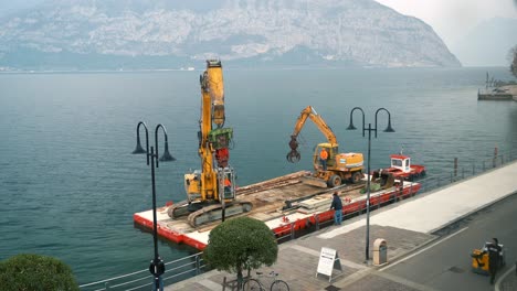 Time-lapse-floating-construction-site,-barge-ship-with-excavator-industrial-equipment-on-lake-Iseo-repairing-shore-promenade