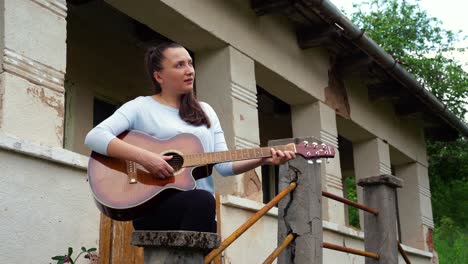 Eine-Junge-Frau-Spielt-Gitarre-In-Der-Nähe-Eines-Alten-Hauses
