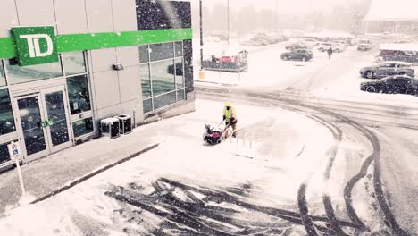 Aerial-drone-shot-of-a-man-clearing-snow-and-ice-with-a-snow-blower-from-the-walkway-in-front-of-a-Toronto-Dominion-Bank-branch-during-a-winter-blizzard,-Ontario,-Canada