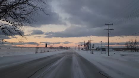 POV-shot-of-a-vehicle-traveling-down-a-slippery-icy-road-after-a-recent-snowstorm,-strong-winds-blowing-ice-across-the-road-as-the-sun-sets-in-the-distance-in-Owen-Sound,-Ontario,-Canada