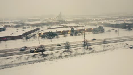 Aerial-drone-shot-tracking-a-snow-plough-truck-traveling-along-a-busy-road-during-a-snow-storm-with-poor-visibility,-Toronto,-Canada