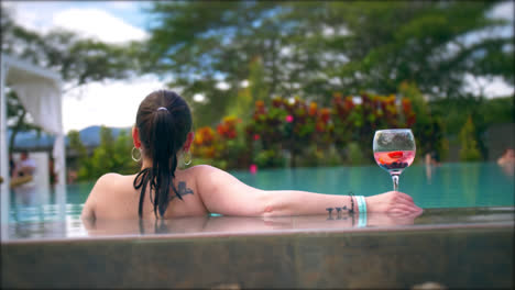 Young-dark-haired-woman-in-swimming-pool-with-drink,-seen-from-behind