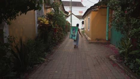 A-female-artist-walking-away-down-a-passageway-between-buildings-holding-an-abstract-painting-over-her-shoulder,-India