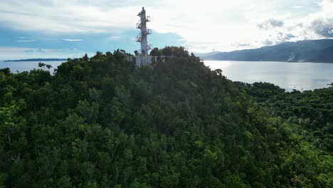 Breathtaking,-Rising-drone-shot-of-Bote-Lighthouse-on-top-of-rainforest-covered-mountains-with-stunning-ocean-bay-and-clouds-in-background