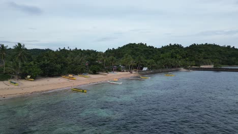 Aerial-Orbit-View-of-idyllic-bangka-boats-lined-against-white-sand-beach-with-stunning-jungle-covered-community-in-the-background