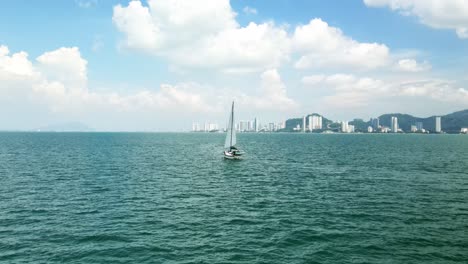 Sailboat-sails-towards-the-Malaysia-Langkawi-Island-Archipelago-with-Mountains,-skyscrapers,-and-tall-buildings-in-the-distance