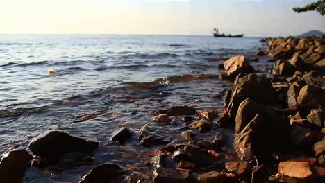 Wellen-Plätschern-An-Einem-Felsigen-Strand-Von-Rabbit-Island-Oder-Koh-Tonsay-In-Kambodscha,-Am-Horizont-Fährt-Ein-Boot-Vorbei-Und-In-Der-Nähe-Schwimmt-Ein-Ablenkender-Plastikbecher,-Der-Das-Ausmaß-Der-Verschmutzung-Zeigt