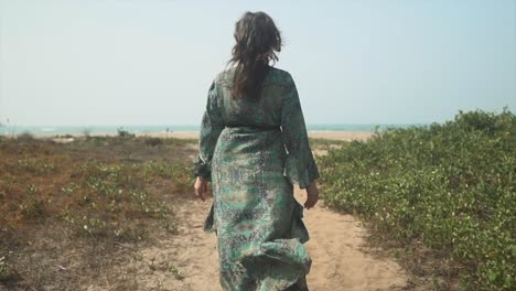 Female-walking-down-path-towards-beach-in-summer-with-long-flowing-dress