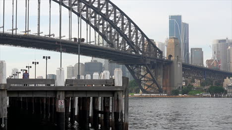 Un-Hombre-Cruza-Un-Paseo-Marítimo-Junto-Al-Puerto-Con-La-Ciudad-De-Sydney-Al-Fondo