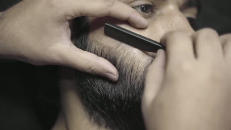 Closeup-Of-Barber-Styling-Mans-Beard-With-Razor-Blade-In-Barbershop