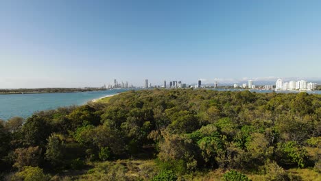 Revealing-drone-view-of-a-sand-Island-covered-with-native-vegetation-close-to-an-urban-city-skyline