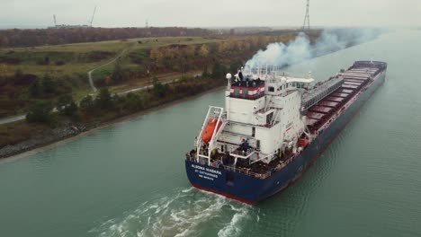 Barge-billows-smoke-transporting-cargo-down-river,-aerial-view-from-behind