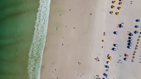 Strand-Mit-Grünem-Blick-Auf-Das-Meer-Von-Oben-Bis-Unten,-Mit-Menschen,-Die-An-Einem-Sommerurlaubstag-Spaß-Haben,-Und-Sonnenschirmen-Und-Farbigen-Sonnenschirmen,-Die-Die-Wellen-Im-Sand-Von-Arraial-Do-Cabo,-Brasilien,-Abdecken
