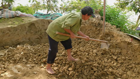 A-side-back-shot-of-an-Asian-man-breaking-clay-soil-with-a-hoe,-using-it-as-raw-material-for-traditional-Thanh-Ha-pottery,-under-the-bright-sun
