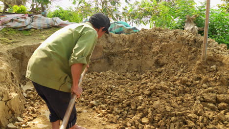 An-Asian-man-breaks-clay-soil-with-a-hoe,-preparing-raw-material-for-traditional-pottery-in-Thanh-Ha-village,-illuminated-by-the-warm-sun