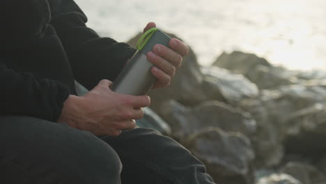 close-up-man-unscrews-thermos-and-pour-hot-drink-into-cup,-then-drinks-it-as-calm-waves-wash-over-rocks-in-background-during-golden-hour
