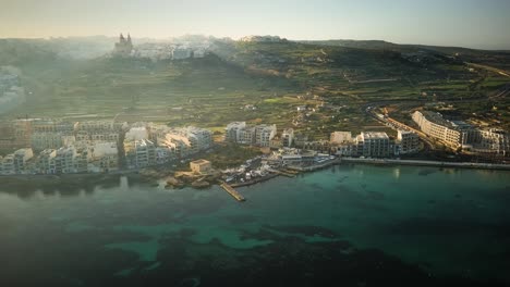 Drone-Orbit-of-Mellieha-bay-from-a-distance-on-a-heavily-misty-morning-in-Malta-with-Mellieha-church-on-hill-in-background